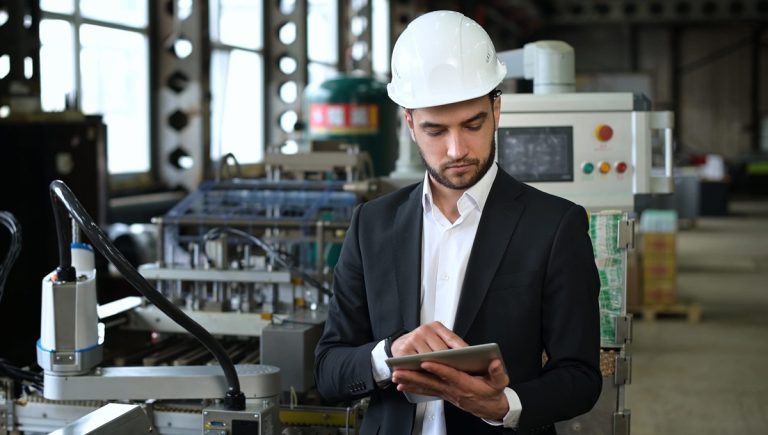 Professionista della sicurezza sul lavoro con caschetto protettivo e tablet mentre verifica i dati tecnici di un macchinario industriale conforme alle norme armonizzate 2026.