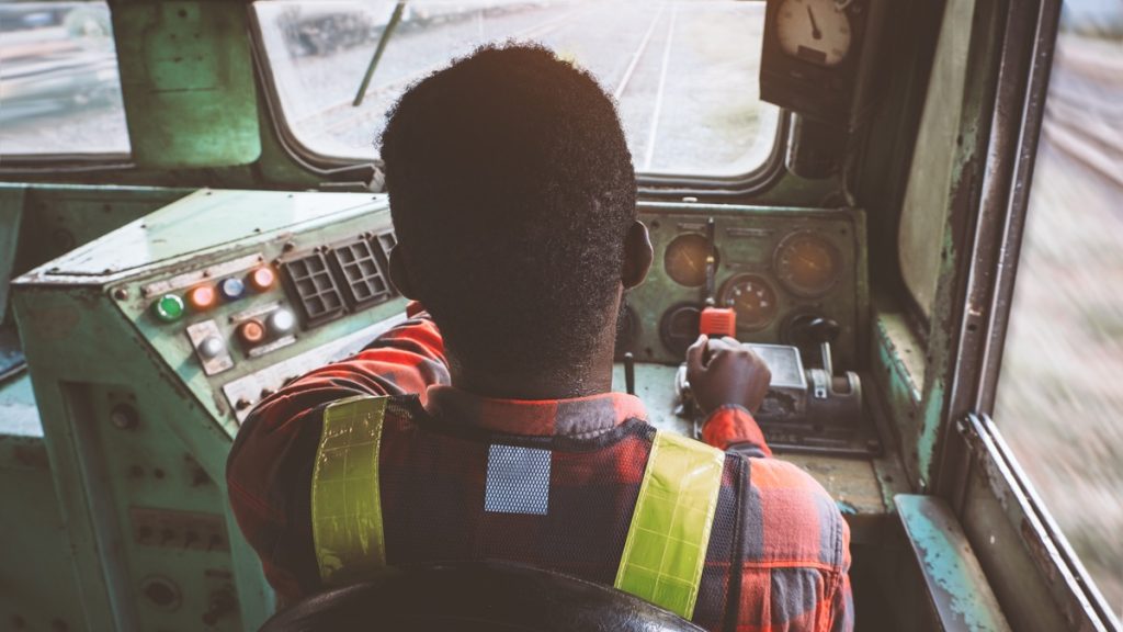 Veduta interna della cabina di guida di un treno con macchinista al lavoro: focus sulla vigilanza e sicurezza ferroviaria.