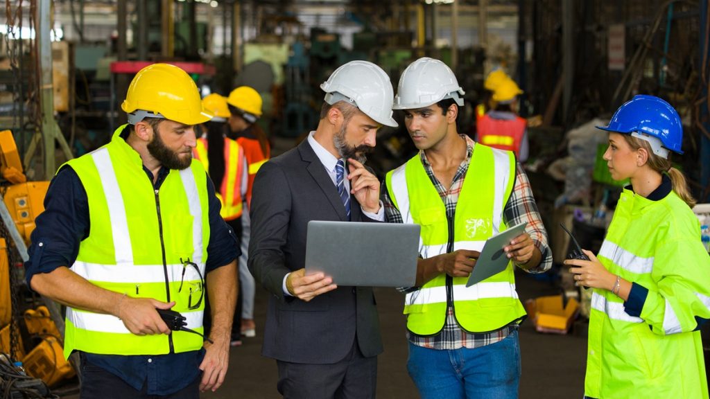 Professionista RSPP in abito scuro con laptop monitora e si confronta con tecnici e operatori.