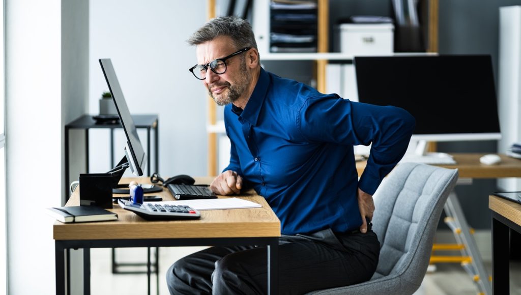 Uomo alla scrivania che soffre di mal di schiena causato da postura scorretta al computer, focus del corso sull'ergonomia.