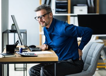 Uomo alla scrivania che soffre di mal di schiena causato da postura scorretta al computer, focus del corso sull'ergonomia.