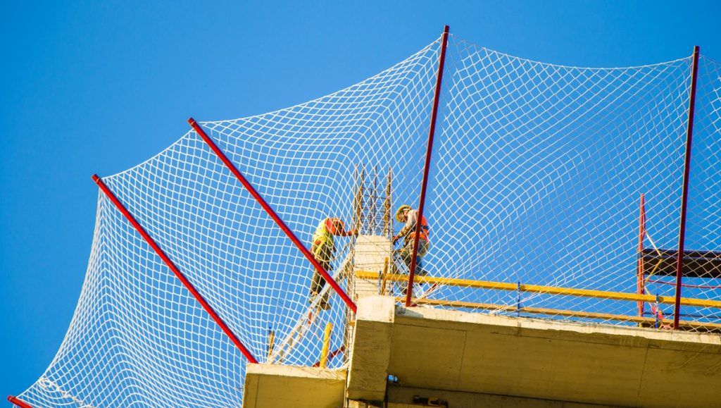 Cantiere edile: installazione di sistema di reti di sicurezza a ventaglio (tipo T) contro la caduta di oggetti e detriti, con cielo azzurro sullo sfondo.