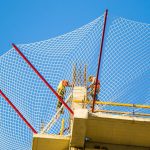 Cantiere edile: installazione di sistema di reti di sicurezza a ventaglio (tipo T) contro la caduta di oggetti e detriti, con cielo azzurro sullo sfondo.