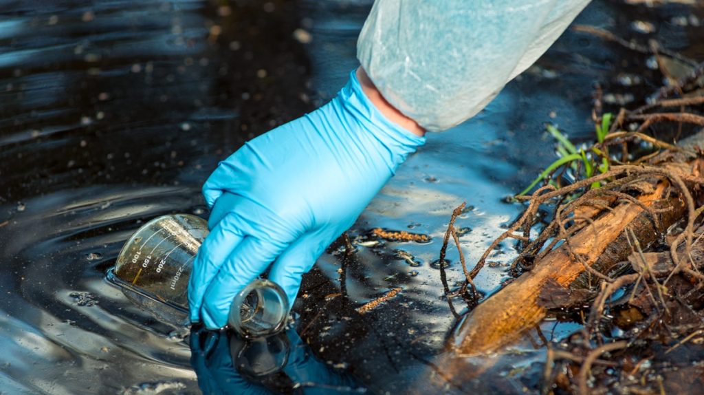 Mano con guanto di nitrile raccoglie campione d'acqua con becher per analisi secondo Direttiva 2026/805